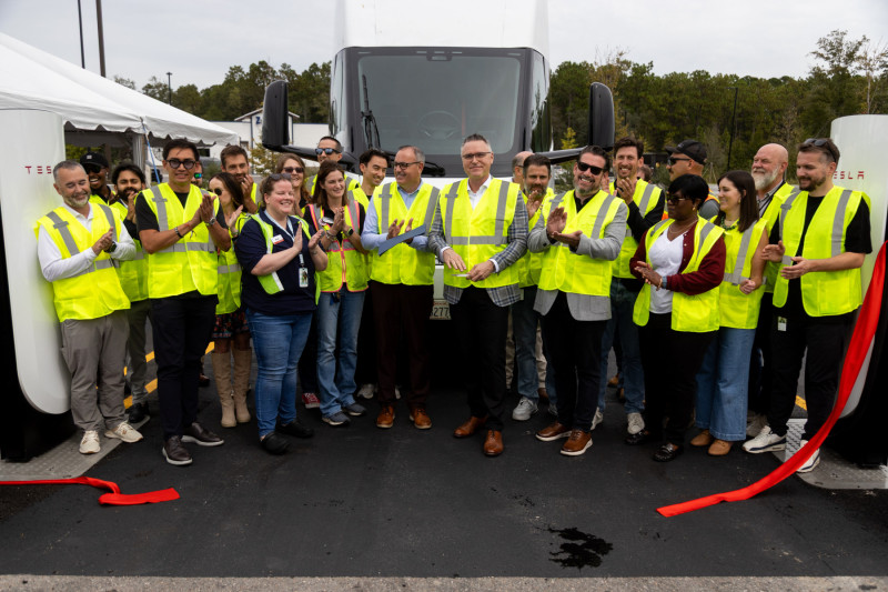 Opening-event attendees holding scissors beside a cut red ribbon between Tesla charging stalls