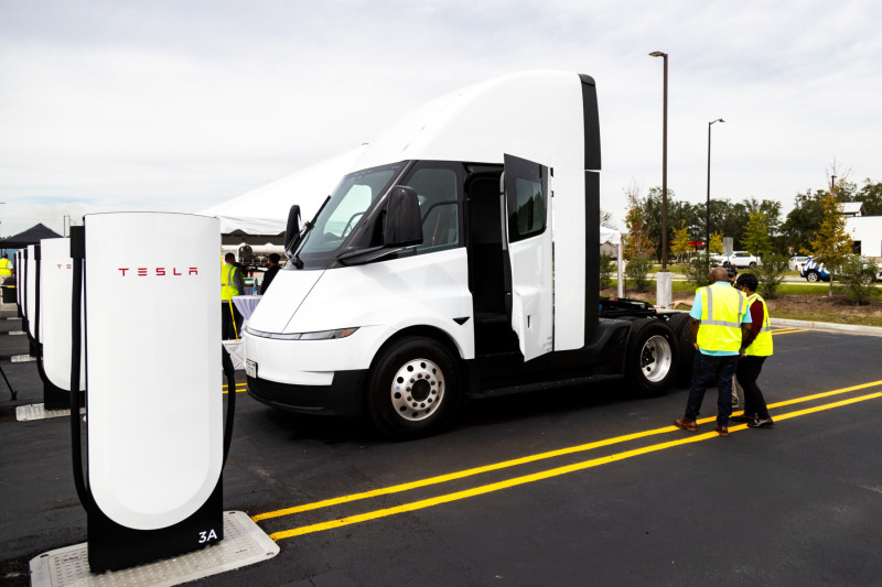 White Tesla Semi beside a Tesla-branded charging stall, with people in high-visibility vests nearby